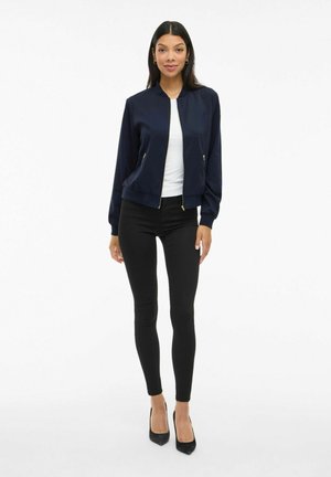 Femme debout, portant une veste bomber marine sur un haut blanc, un pantalon skinny noir et des escarpins noirs sur fond blanc.