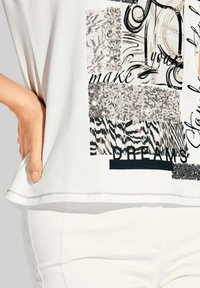 Witte t-shirt met een collage van zwarte en zilveren patronen, waaronder zebrapatronen en pailletten, met de tekst "vervul je dromen."