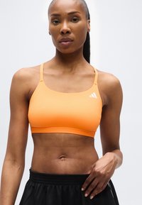 Γυναίκα που φοράει πορτοκαλί αθλητικό σουτιέν και μαύρο σορτς, στέκεται μπροστά σε ένα απλό φωτεινό φόντο, με το ένα χέρι να αγγίζει το σορτς της.