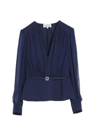 Kvinde iført navyblå gennemsigtig bluse med ærmer og bukser, holder en sølv clutch, har guld stroppe høje hæle på, vender fronten mod kameraet.