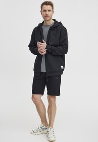 Čierna mikina na zips so šedým tričkom s kruhovým výstrihom, doplnená čiernymi šortkami. Medzi vlastnosťami je logo na hrudi a športová obuv svetlých farieb.