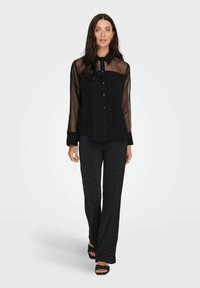 Crna košulja na dugih rukava s prozirnim rukavima, uparen s crnim hlačama koje imaju bijele točkice. Cipele s ravnim crnim potplatima upotpunjuju odjevnu kombinaciju.