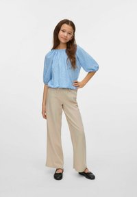 Lichtblauwe blouse met een klein wit bloemenpatroon, ballonslouders, gecombineerd met beige wijde broekspijpen en zwarte platte schoenen.