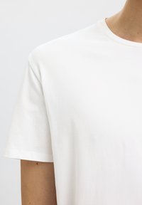 Gros plan d'une personne portant un t-shirt blanc uni à manches courtes avec un col rond sur un fond clair.