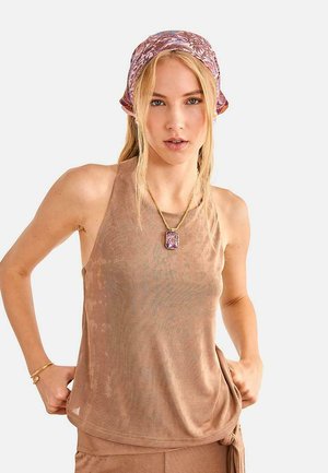 Mujer rubia con un top marrón sin mangas, pantalones a juego, un pañuelo morado con estampado y un collar con colgante morado, posando con las manos en las caderas.