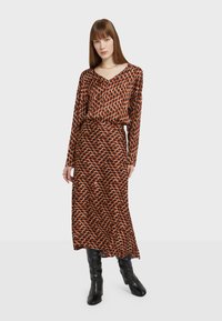 Pitkähihaisen maxi-mekon väri on ruskea, ja siinä on oransseja ja kermaisia geometrisia kuvioita. Mekossa on V-pääntie ja vyötäröllä on vyö, joka on yhdistetty mustiin saappaisiin.