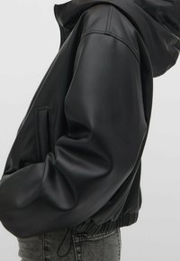 Veste noire en similicuir avec capuche. Présente une coupe ample, une taille élastique et une poche latérale. Texture lisse avec des détails de coutures visibles.