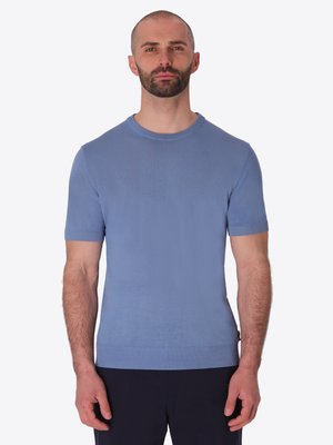 Uomo con barba corta che indossa una camicia a maglia azzurro chiaro aderente a maniche corte e pantaloni scuri, in piedi davanti a uno sfondo semplice.