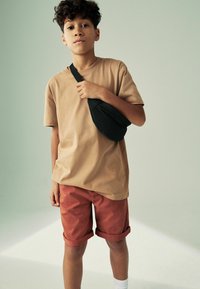 T-shirt en coton marron, manches rollées, associé à un short rouge à revers. Sac à bandoulière noir porté en travers du corps, chaussettes blanches, arrière-plan neutre.