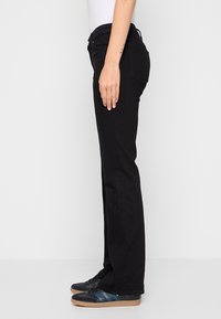 Vue latérale d'une personne portant un jean noir droit, des baskets noires et bleues, et un haut blanc avec un bras relâché le long du corps.