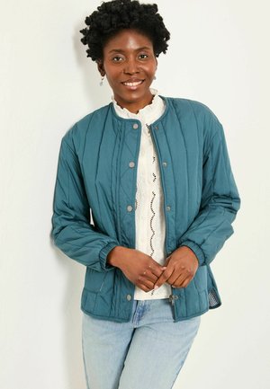Teal quilted jacket with a round neckline, snap buttons, and elasticized cuffs. Paired with a white textured blouse and light blue jeans.