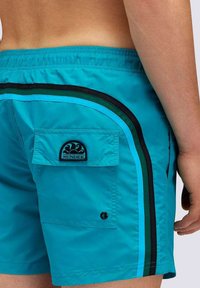 Pantaloni da bagno turchesi con strisce laterali nere e blu, dotati di una tasca sul retro e un'etichetta con logo. Realizzati in materiale leggero e ad asciugatura rapida.