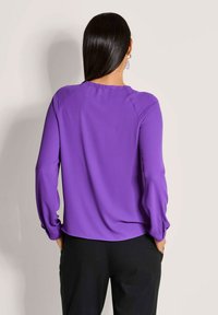 Paarse blouse met lange mouwen van gladde stof. Heeft een ronde halslijn, details met geplooide manchetten en een relaxte pasvorm. Gekleed met zwarte broeken.