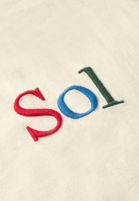 Bílá látka s vyšitým nápisem "Sol" v červené, modré a zelené nitě. Písmena mají texturu a jsou mírně vystouplá. Jednoduchý a jasný design.