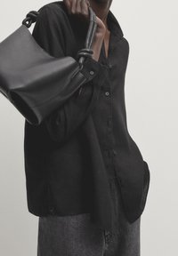 Chemise noire en lin à boutons avec une coupe décontractée, assortie à un pantalon en denim foncé. Un sac en cuir noir avec une poignée en corde texturée est tenu en main.
