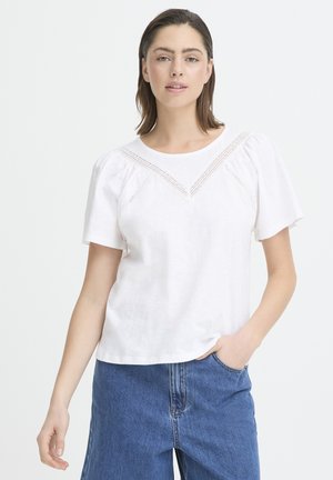 Junge Frau mit schulterlangem braunem Haar, die eine weiße Kurzarmbluse mit Spitzendetails und blaue Jeans trägt, eine Hand in der Tasche.