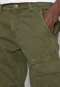 Olivgrön cargobyxor tillverkade av slitstarkt tyg, med sidofickor, knappstängning och synliga söm detaljer.