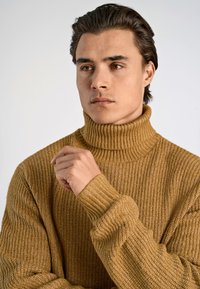 Beige ribbad tröja med hög, vikt krage. Stickad struktur är grov, med långa ärmar och en avslappnad passform.