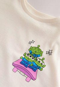 White t-shirt with a graphic of three green alien figures on a pink platform, wearing blue outfits, surrounded by small stars.