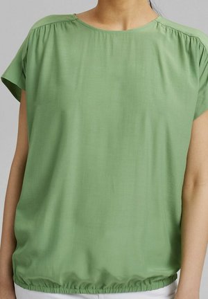 Kobieta ubrana w luźną, zieloną bluzkę z krótkim rękawem, marszczonymi ramionami i elastycznym dołem, zestawioną z białymi spodniami.