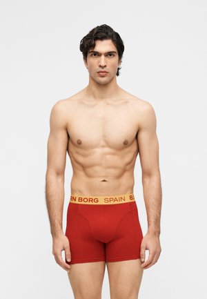 Mez nélküli, rövid göndör sötét hajú férfi piros alsónadrágot visel, amelynek sárga derékrésze „BORG SPAIN” felirattal rendelkezik, egy sima háttér előtt áll.