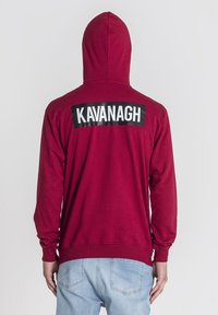 Sudadera con capucha color marrón burdeos y mangas largas, que presenta una etiqueta negra rectangular en la parte de atrás con el texto blanco "KAVANAGH". Material de mezcla de algodón.