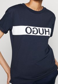 Mørkeblå bomulds t-shirt med opbrettede korte ærmer, forsynet med en hvid vandret stribe, der viser ordet "HUGO" i fede sorte bogstaver.