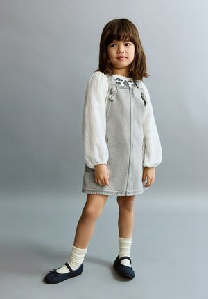 Jeune fille aux cheveux bruns portant un chemisier blanc à manches longues, une robe grise, des chaussettes blanches et des chaussures bleu marine, debout devant un fond gris.