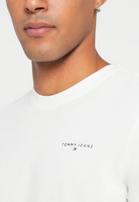 Fehér pamut póló kerek nyakkal és rövid ujjakkal, fekete "TOMMY JEANS" logóval a bal mellen.