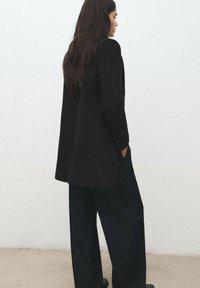 Długa czarna marynarka wykonana z teksturowanej tkaniny, o prostym kroju i pojedynczym guziku. Połączona z szerokimi spodniami z ciemnego denimu.