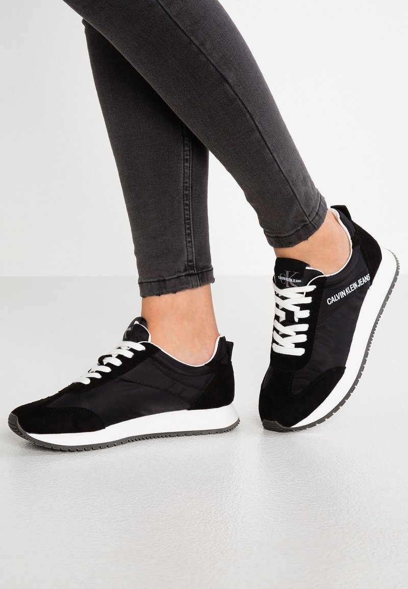 Pieds portant des baskets Calvin Klein noires et blanches avec des lacets blancs et le logo Calvin Klein Jeans sur les côtés, associés à un jean skinny gris foncé.