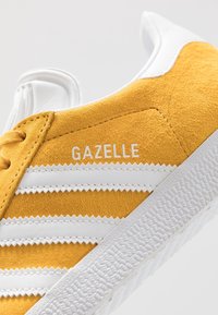 Gul mockaskinsneaker med vita läderdetaljer, med tre vita ränder och ordet "GAZELLE" tryckt i vitt. Texturerad gummisula.