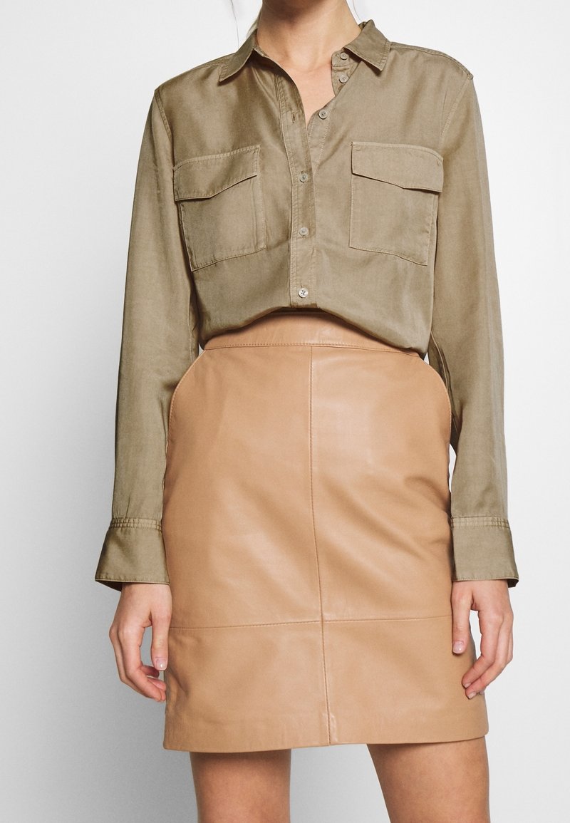 Femme portant une chemise boutonnée vert olive avec des poches à rabat et une jupe en cuir beige taille haute, debout devant un fond blanc.