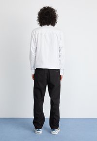 Weiß gestreiftes Langarmhemd mit Knopfmanschetten, schwarze Hose und gemusterte Sneaker, von hinten vor einem schlichten Hintergrund betrachtet.