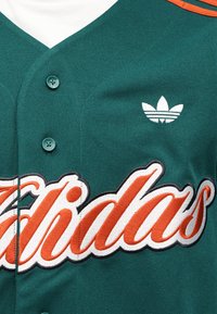 Sinine-roheline spordisärk oranži tikitud "Adidas" logoga. Omadustega V-kaelus, nööpidega sulgemine ja väike valge Adidas logo rinnal.