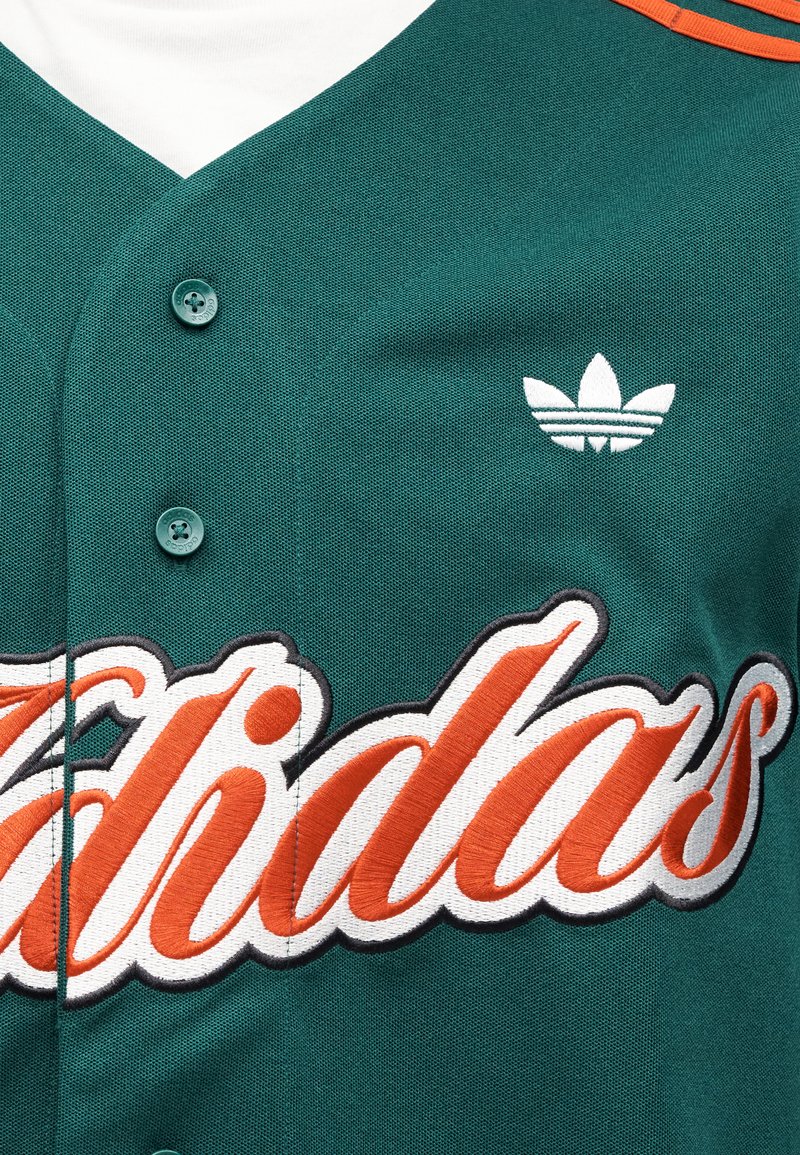 Maillot de sport bleu sarcelle avec logo "Adidas" brodé en orange. Comprend un col en V, une fermeture à boutons, et un petit logo Adidas blanc sur la poitrine.