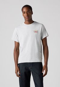 Grijze katoenen T-shirt met korte mouwen, met een kleurrijk bedrukt logo op de linkerborst. Gecombineerd met een donkere spijkerbroek.