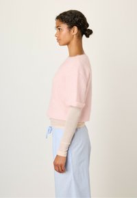 Lichtroze, cropped trui met korte mouwen, gemaakt van zachte stof. Gelaagd over een beige lange mouwen top en gecombineerd met lichtblauwe gestreepte broek.