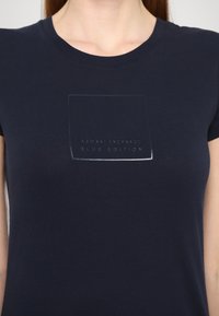 Námořnická modrá pánská polotriko s krátkým rukávem vyrobené z bavlny, s texturovaným logem ve tonálním rámečku, na kterém je uprostřed nápis "Armani Exchange Blue Edition".