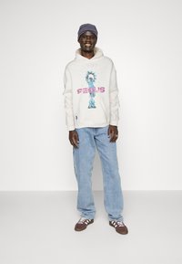 Sudadera crema con estampado azul y texto rosa, pantalones vaqueros holgados color azul claro, y zapatillas marrones con rayas blancas. Gorro de punto con patrón usado en la cabeza.