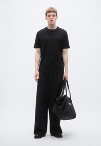 Fekete rövid ujjú póló domború "BOSS" logóval, fekete bő szárú nadrággal és hozzá illő fekete táskával, amelyen fehér szöveg látható.