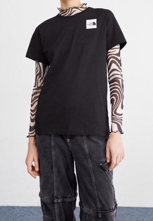 Personne portant un t-shirt noir The North Face par-dessus un top transparent à manches longues imprimé zèbre et un pantalon cargo noir ample, debout devant un fond uni.