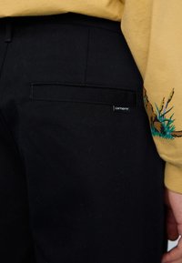 Zwarte geweven broek met een zijzak, met een subtiele logo-tag. Een gele sweatshirt met een kleurrijke grafische print is gedeeltelijk zichtbaar.
