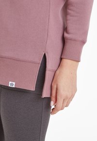 Dusty pink sweatshirt with ribbed cuffs, side slits, subtle texture, and a logo tag at the hem, paired with dark leggings.
