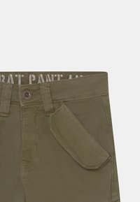 Olivgröna byxor i texturerat tyg med knapp framtill, synlig sömnad och sidofickdesign. Etiketten visar "PANTALON."