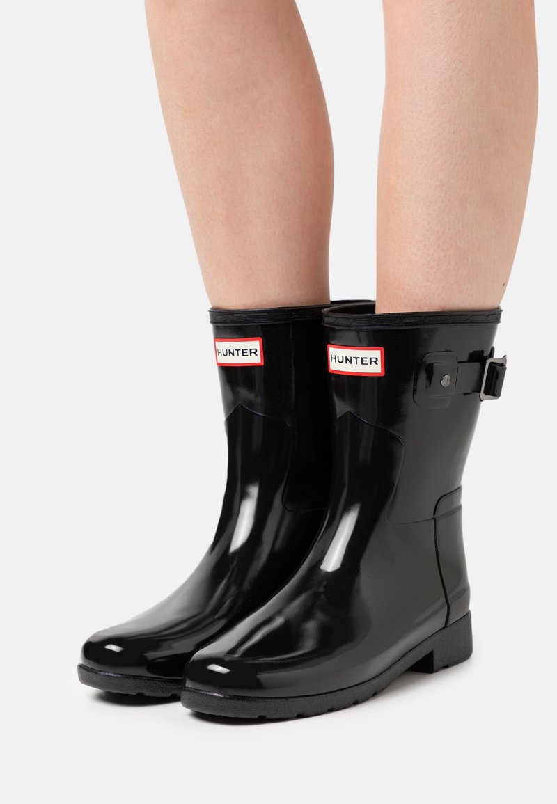 Bottes de pluie en caoutchouc noir avec une finition brillante, présentant une étiquette de marque rouge et blanche "HUNTER" à l'avant et un détail de boucle réglable sur le côté.