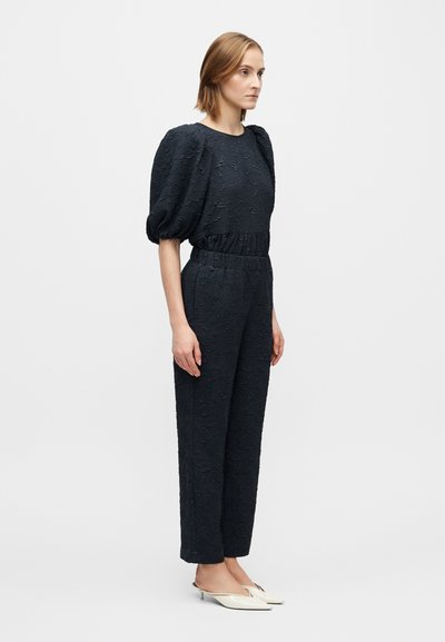 Combinezon bleumarin cu model floral texturat, mâneci bufante, talie strânsă și picioare conice, asortat cu pantofi albi slip-on.