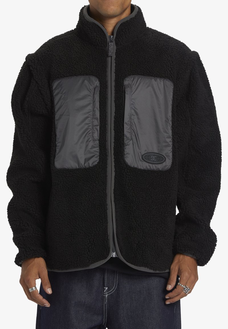 Svart fleecejacka med grå nylon detaljer och dragkedjefickor. Har en hög krage och elastiska ärmslut. Mjuk textur och avslappnad passform.