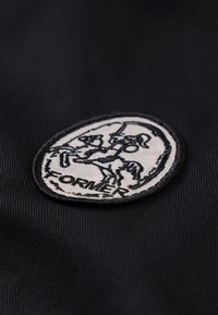 Μαύρο ύφασμα με κυκλικό κεντητό έμβλημα σε λευκό φόντο, μαύρο περίγραμμα και γραφικό με στυλιζαρισμένη φιγούρα και το κείμενο «FORMER».