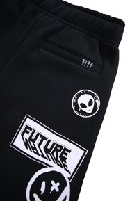 Pantalons de survêtement noirs avec taille élastique, ornés de motifs graphiques blancs : une tête d'extraterrestre, le texte "FUTURE" et un motif de visage souriant.